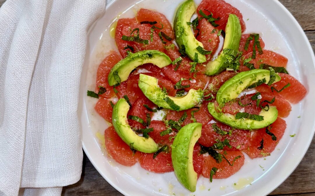 Grapefruit and Avocado Salad. Sliced grapefruit with sliced avocado arranged on a white plate garnished with lime juice, lime zest, salt, and a little drizzle of olive oil with a white napkin beside the plate.