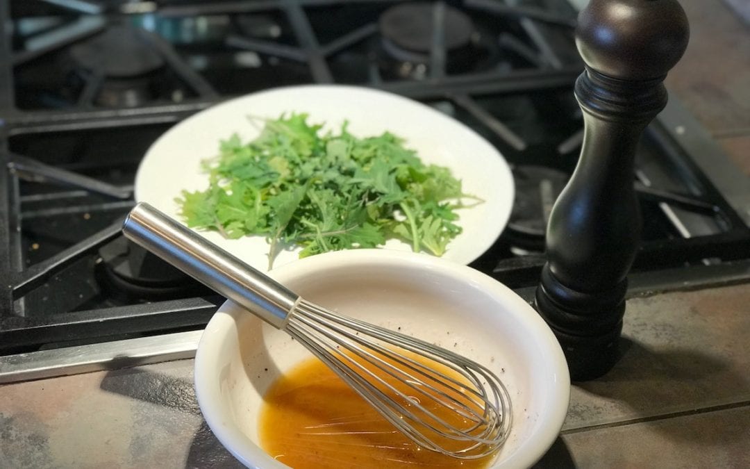 Class red wine vinaigrette. White bowl with red wine vinaigrette whisk and a plate of arugula sitting behind it.