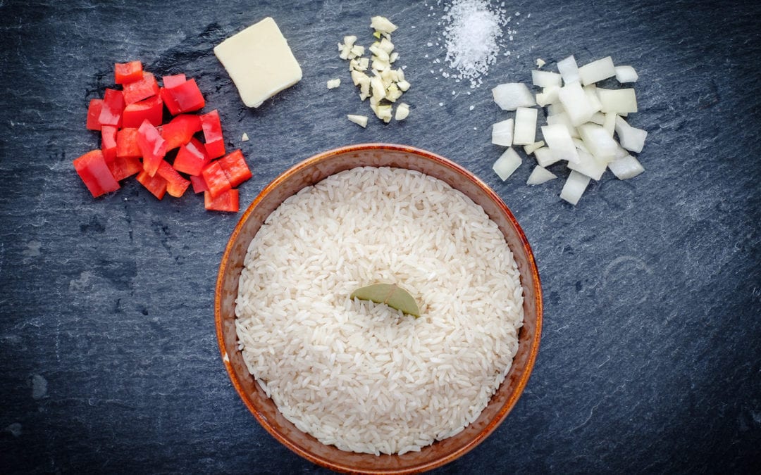 How To Make Rice. Raw ingredients to make rice, including rice in a bowl surrounded by red bell pepper, a pat of butter, garlic, salt, and onion. a bay leaf decorates the rice