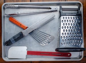A fish spatula, box grater, silicon spatula, tong and microplane on top of an aluminum tray on the table.