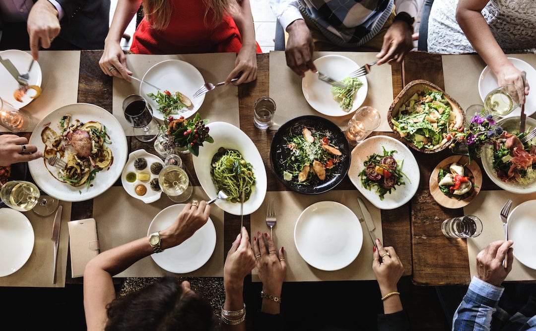 The secret to health and happy is Life At The Table. An overhead shot of a long table set with dinner with several friends and family enjoying dinner.