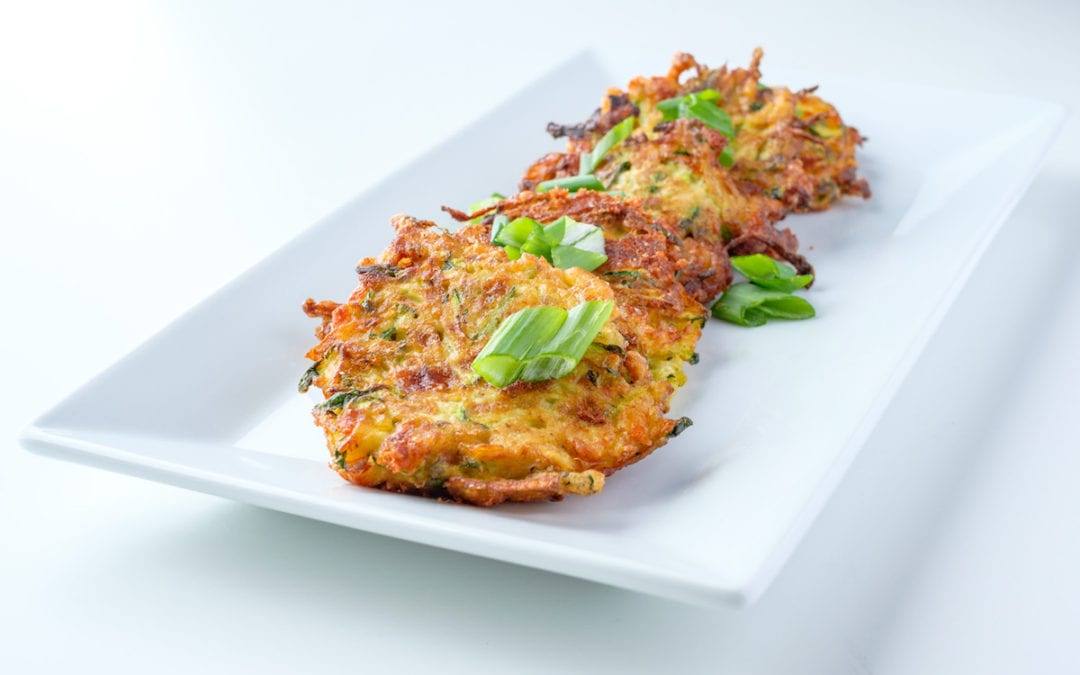 Zucchini Fritters. Several patties of zucchini fritters, garnished with scallion slices on a white plate.