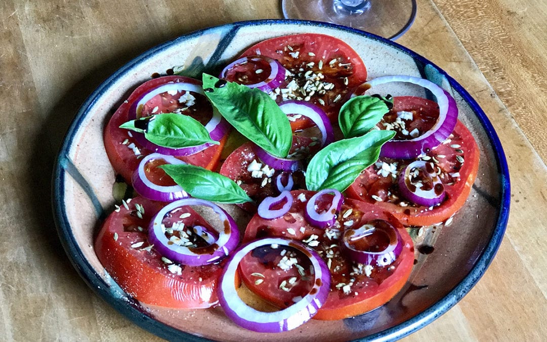 Slices of fresh tomatoes with slices of fresh onions with a dash of soy sauce and a sprinkle of anise seeds topped with cilantro leaves on a plate on top of the table.