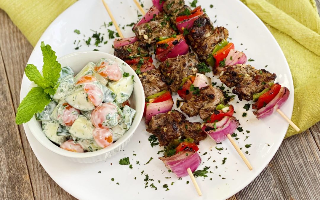 Life At The Table Shish Kebab. Three shish kebab skewers on a white plate garnished with parsley with a bowl of tomato, cucumber, and yogurt salad.