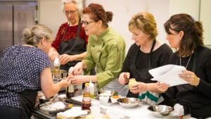 What's for dinner? Life At The Table. Chef Sandra in a green chef jacket assisting a student in a cooking class with several other students gathered around. The class is making a vinaigrette.