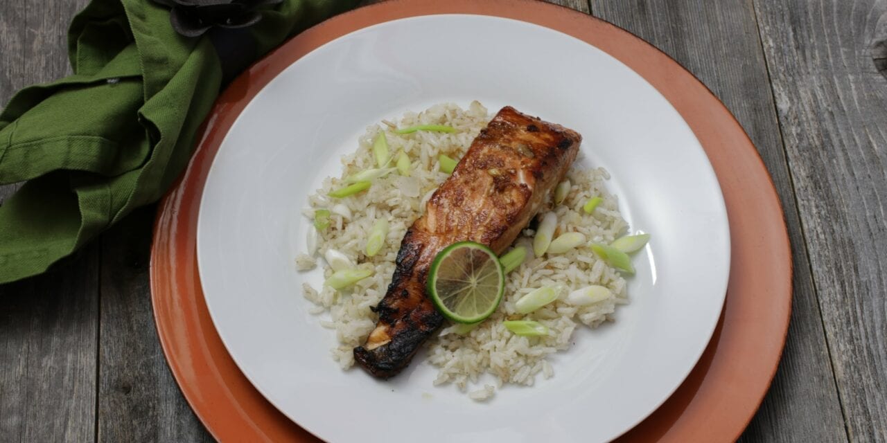 Pan-seared salmon that has been marinated in the Cowlitz River Salmon Marinade. It's presented on a white plate with an orange charger underneath served over cardamom rice and garnished with a lime.