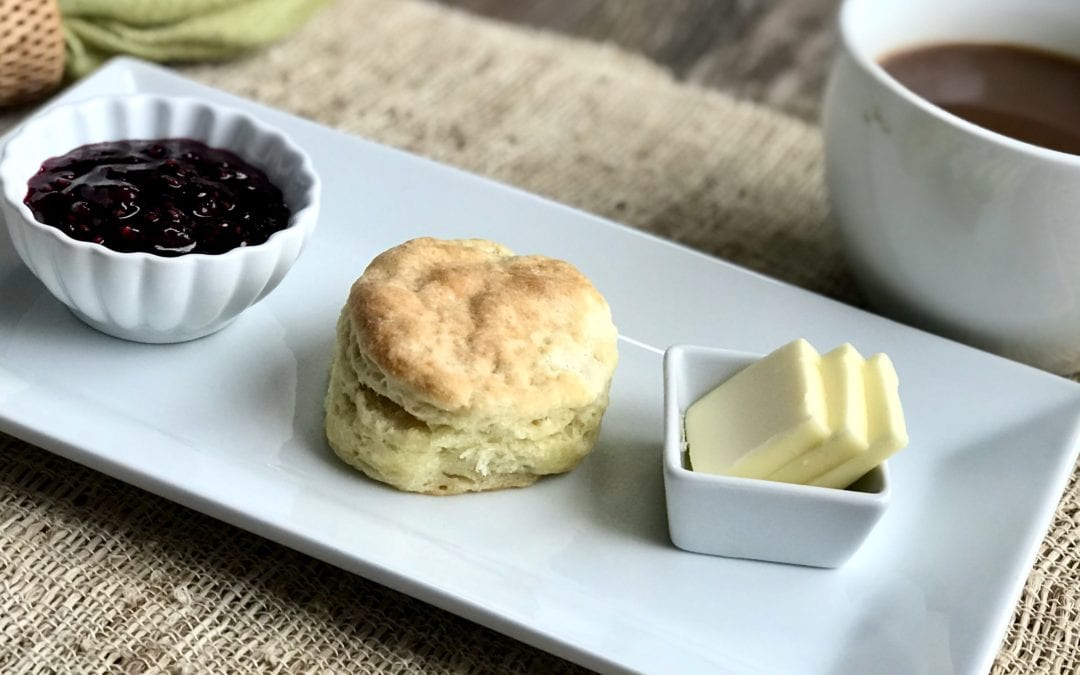 Life At The Table | A Homemade Biscuit Recipe Fit For a King. A single biscuit on a white platter with several pats of butter and a small bowl of jam on either side. The platter is sitting on a woven placemat with a cup of coffee and a green napkin on either side.