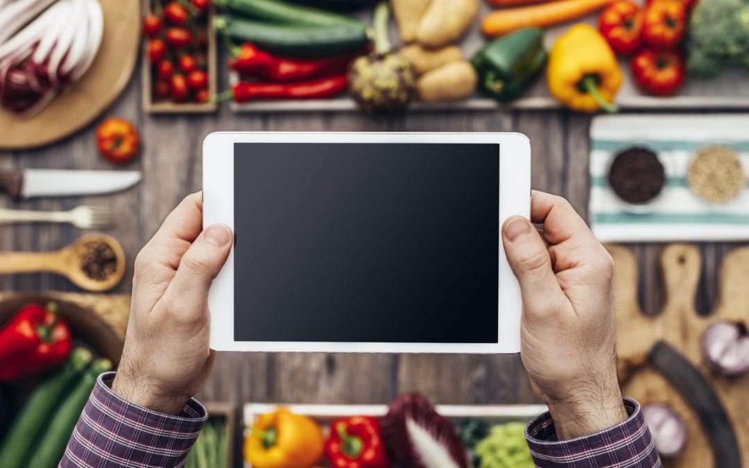 Virtual, Live Cooking Classes | Life At The Table. Top down view of a man holding an iPad over a cutting board with vegetables, bell peppers, radicchio, mushrooms, and broccoli.