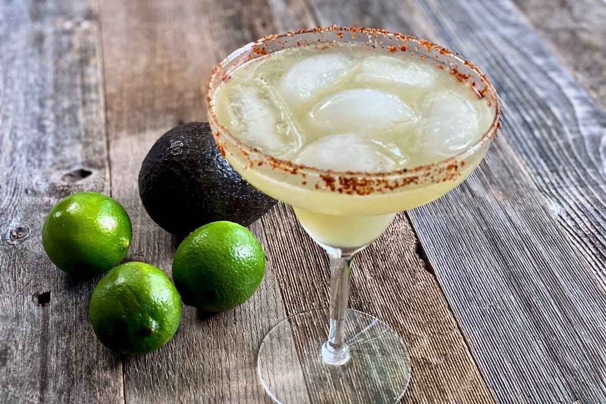 Margarita glass filled with a fresh lime juice margarita sitting on a weathered table with limes and an avocado next to it.