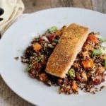 Life At The Table Mediterranean Quinoa Salad Virtual Cooking Class - A slice of seared salmon on a bed of quinoa and vegetables. The meal is on a round, white plate sitting on a wooden table along with a beige, cloth napkin