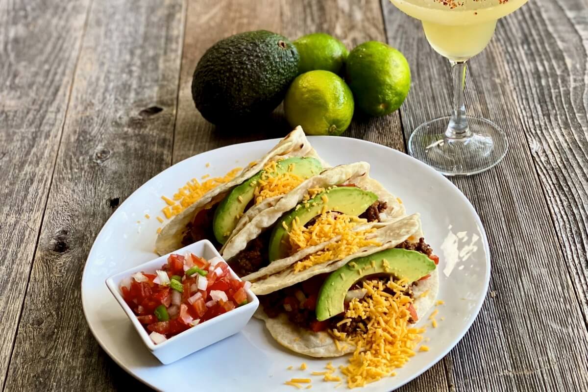 Life At The Table Virtual Culinary Team Building Events.Three homemade tacos on a white plate with avocado, cheese, and fresh salsa with a margarita sitting next to the plate on a wooden table top.