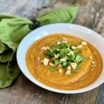 A delicious bowl of butternut squash soup in a white bowl, garnished with cilantro and chopped peanuts, on a wooden table. Butternut Squash Soup: Life At The Table