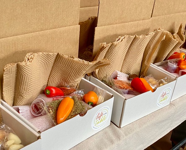 Life At The Table Ingredient Kits Small, white, rectangular, cardboard boxes lined up on a table. Mini bell peppers, packets of Italian seasoning, and small plastic cups of red wine vinegar can be seen in the boxes