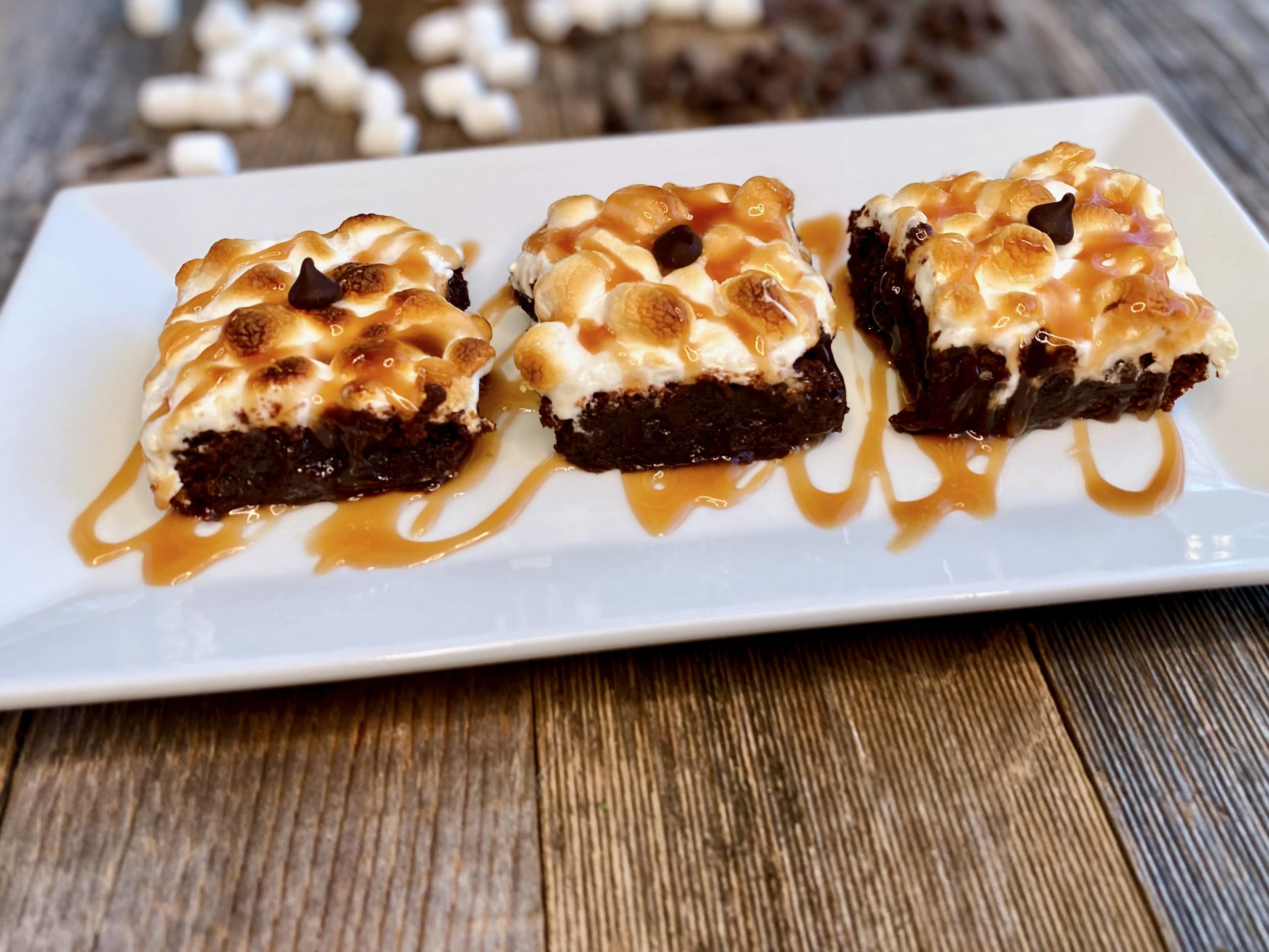 Life At The Table Decadent Brownies | Three brownies on a white platter with toasted marshmallow and drizzled with salted caramel sauce.