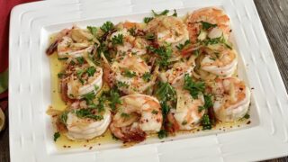 Life At The Table Shrimp al Ajillo Happy Hour Event. Shrimp al ajillo on a white plate garnished with parsley sitting on a wooden table with a red napkin beside it.