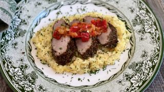 Life At The Table Jolly Juniper Pork Tenderloin Culinary Team Building. Juniper berry pork tenderloin, sliced on top of polenta and garnished with a tomato jam on a festive plate sitting on a wooden table.