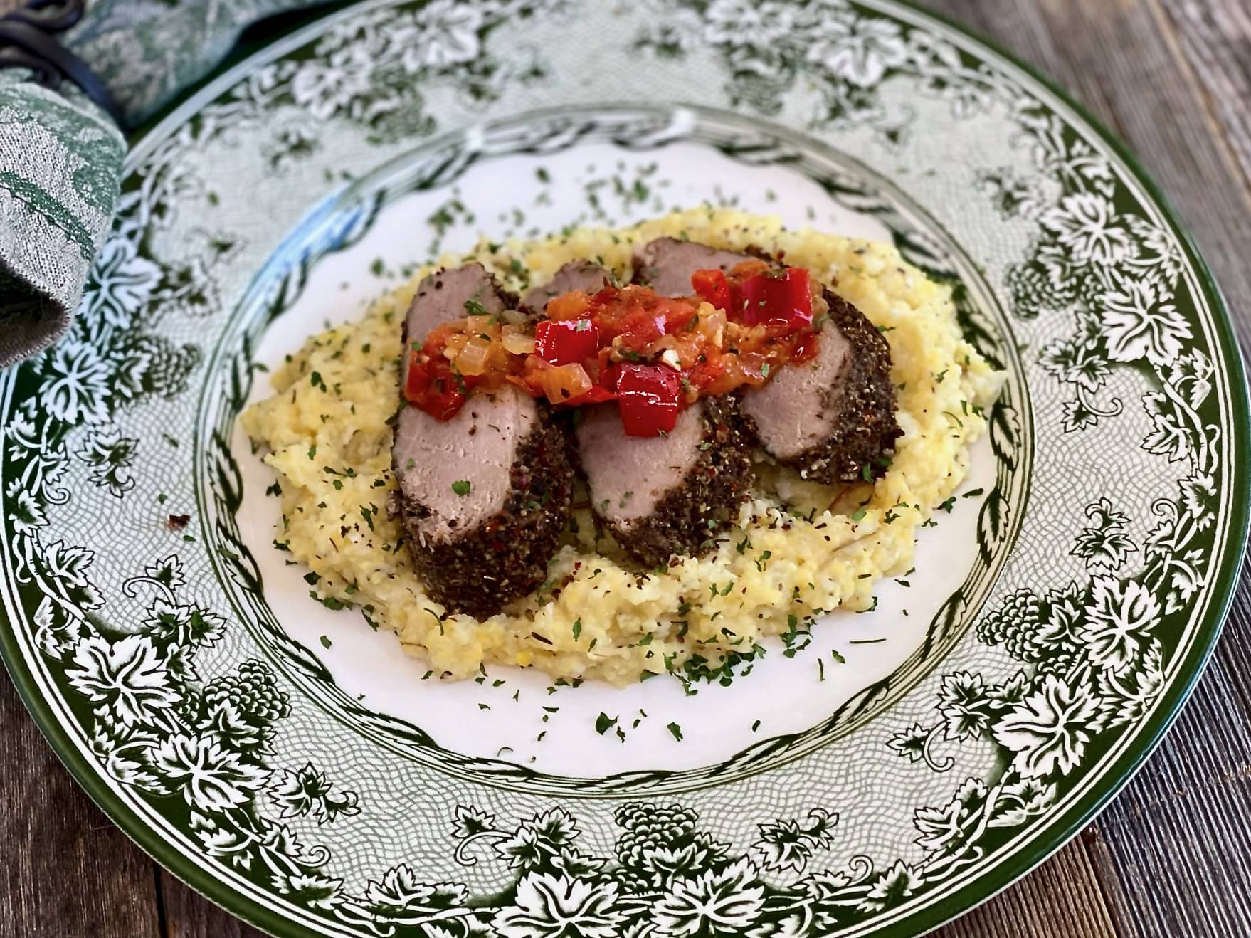 Life At The Table Virtual Culinary Team Building Events. Jolly Juniper Roasted Pork Tenderloin with Chef Sandra's Jolly Juniper seasoning sliced on a plate sitting on a bed of parmesan grits on a green leafy plate with a napkin beside it.