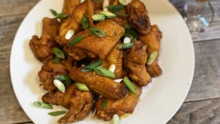 A pile of hot wings on a white plate garnished with green onion sitting on top of a wooden table.