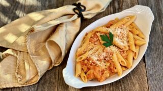 Life At The Table Pasta alla Penne. A white dish of pasta alla penne on a wooden table with a yellow napkin with sitting beside it.