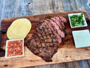 Life At The Table Arrachera | Grilled Arrachera (skirt steak) on a wooden serving platter with corn tortillas, salsa, cilantro, and minced onion.