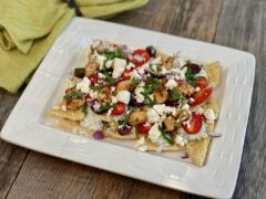 White plate of nachos with chicken, feta cheese, tomatoes, cucumber, onion, and spices sitting on a table next to a green napkin