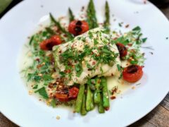 Life At The Table | Baked Cod A white plate with baked cod lying on a bed of asparagus with blistered tomatoes and garnished with fresh herbs.