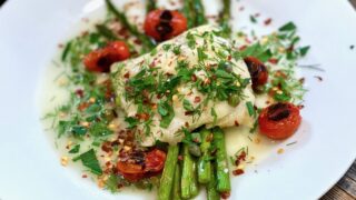 Life At The Table | Baked Cod A white plate with baked cod lying on a bed of asparagus with blistered tomatoes and garnished with fresh herbs.