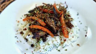 Vaca Frita | Life At The Table. Vaca frita with red bell peppers and onion over a bed of white rice on a white plate sitting on a wicker charger.