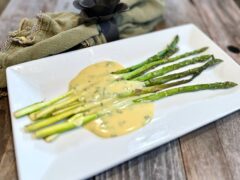Life At The Table | Bêarnaise Sauce. A white plate with roasted asparagus ladled with bêarnaise sauce sitting on a wooden table with a green napkin with a black napkin ring beside it.