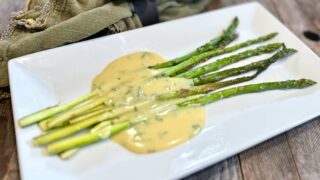 Life At The Table | Bêarnaise Sauce. A white plate with roasted asparagus ladled with bêarnaise sauce sitting on a wooden table with a green napkin with a black napkin ring beside it.