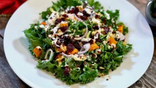 Life At The Table Kale Salad with Creamy Yogurt and Lemon Dressing | kale salad on a white plate with cubed sweet potato, dried cranberries, shallots, and pistachios with a creamy yogurt and lemon dressing on a wooden table with a red napkin beside it.