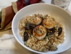 How To Make Risotto | Life At The Table. A bowl of risotto topped with sautéed mushrooms and scallops sitting on a wooden table with a napkin and a glass of wine.