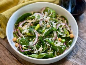 Life At The Table Spinach Salad with a Fennel Seed Dressing. A spinach salad with red onion, parmesan, chickpeas, asparagus, and dill in a white bowl sitting on a wooden table.