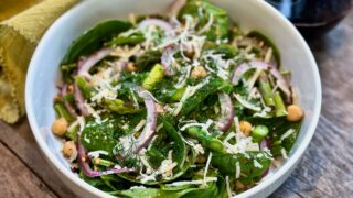 Life At The Table Spinach Salad with a Fennel Seed Dressing. A spinach salad with red onion, parmesan, chickpeas, asparagus, and dill in a white bowl sitting on a wooden table.