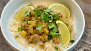 Life At The Table Fiji Chicken Curry. A bowl of Fiji Chicken Curry served on a bed of jasmine rice with fresh limes as a garnish in a white bowl sitting on a wooden table.
