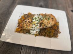 Life At The Table Lentil Salad with Ladolemono. A pan-seared chicken breast sitting on top of a lentil salad with ladolemono. The plate is white and is sitting on a wooden table.