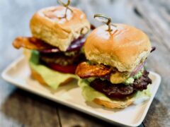 Life At The Table | Smash Burger Sliders. Two smash burger sliders dressed with lettuce, tomato, red onion, avocado, pickle, bacon, and a spicy mango sauce sitting on a white plate on a wooden table.