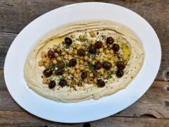 Life At The Table Hummus. Large white plate of hummus garnished with nicoise olives and chick peas on wooden table.