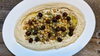 Life At The Table Hummus. Large white plate of hummus garnished with nicoise olives and chick peas on wooden table.