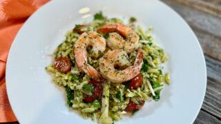 Life At The Table Orzo Salad with Basil Vinaigrette and Poached Shrimp. A white plate with orzo salad with basil vinaigrette with three shrimp on top sitting on a wooden table with an orange napkin beside it.