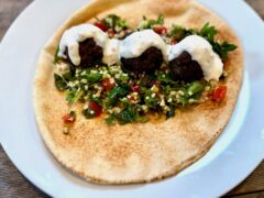 Life At The Table | Kofta (Lebanese Meatballs) A kofta pita sandwich with tabbouleh and hummus on a white plate garnished with a tahini sauce.