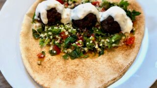 Life At The Table | Kofta (Lebanese Meatballs) A kofta pita sandwich with tabbouleh and hummus on a white plate garnished with a tahini sauce.