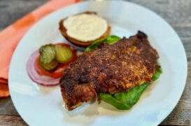 Life At The Table Nashville Hot Chicken Sandwich. A Nashville Hot Chicken Sandwich with a garlic sauce, tomato, lettuce, and red onion on a white plate with an orange napkin beside it sitting on a wooden table.