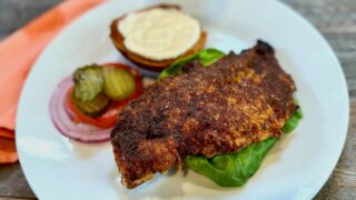 Life At The Table Nashville Hot Chicken Sandwich. A Nashville Hot Chicken Sandwich with a garlic sauce, tomato, lettuce, and red onion on a white plate with an orange napkin beside it sitting on a wooden table.