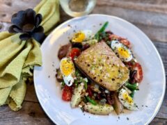 Life At The Table Tuna Niçoise Salad. Tuna Niçoise on a white plate sitting on a wooden table with a green napkin and a glass of white wine.