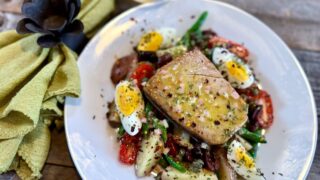 Life At The Table Tuna Niçoise Salad. Tuna Niçoise on a white plate sitting on a wooden table with a green napkin and a glass of white wine.