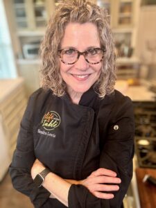 Life At The Table Chef Sandra Lewis in a black chef jacket standing in her home kitchen with her arms crossed looking into the camera.