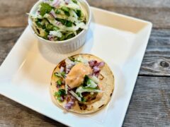 Life At The Table Grilled Shrimp Taco with Chipotle Lime Crema. A street-sized taco on a white plate topped with grilled shrimp, jicama and lime slaw, minced red onion, and a chipotle lime crema.