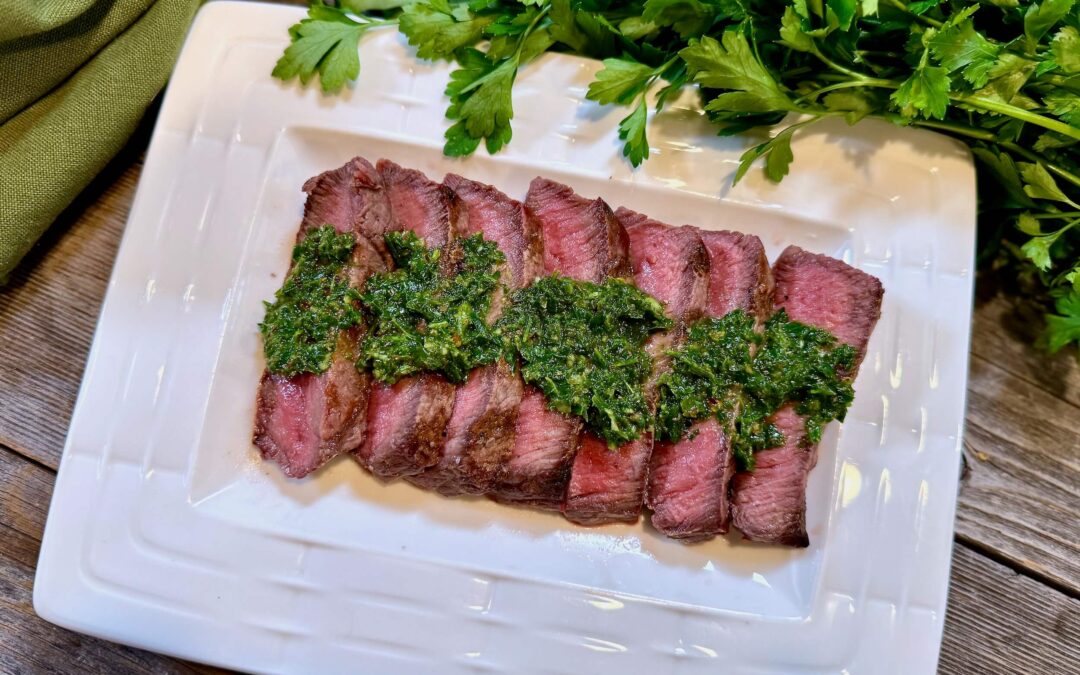 Life At The Table Chimichurri. A flat iron steak drizzled with chimichurri sauce on a white plate sitting on a wooden table with fresh parsley and a green napkin beside it.