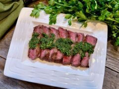 Life At The Table Chimichurri. A flat iron steak drizzled with chimichurri sauce on a white plate sitting on a wooden table with fresh parsley and a green napkin beside it.